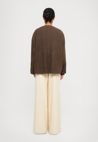 Veste en daim brun, ample, à manches longues, portée sur un pantalon large crème, vue de dos sur un fond blanc.