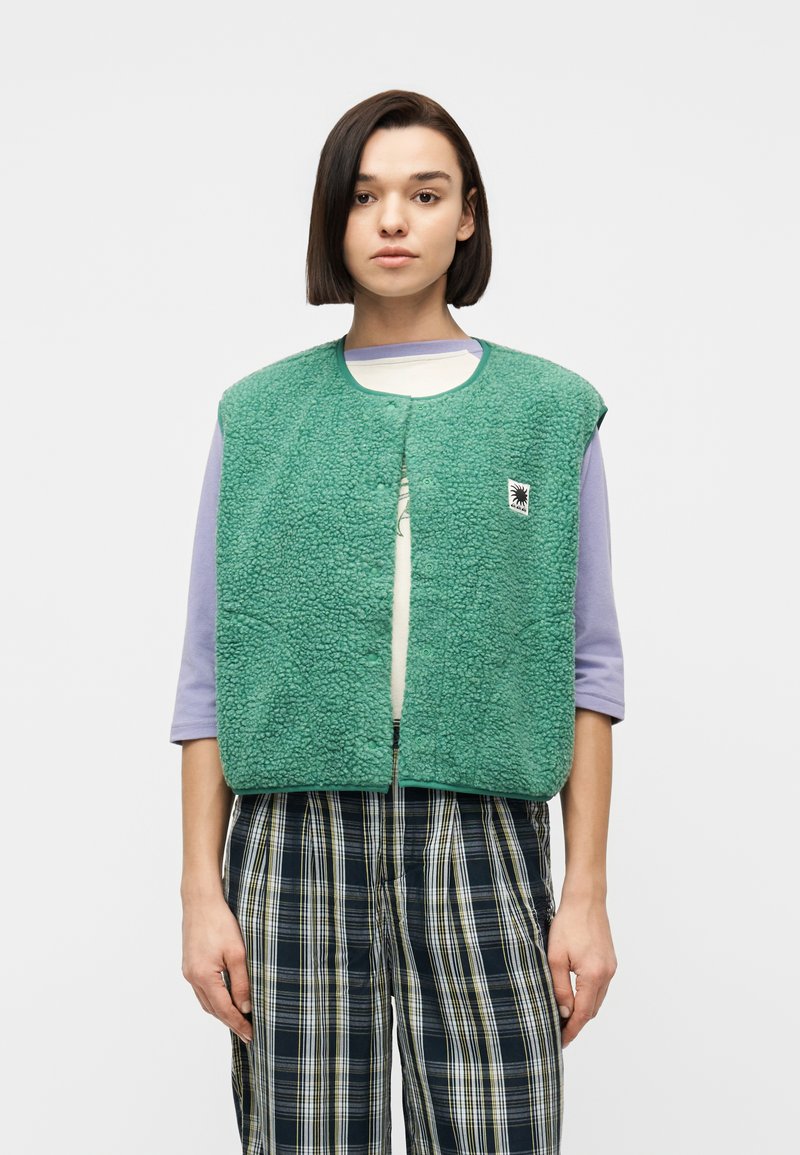 Νεαρή γυναίκα με ίσια σκούρα μαλλιά φοράει πράσινο υφασμάτινο γιλέκο πάνω από πορφυρό μακρυμάνικο πουκάμισο και καρό παντελόνι, στέκεται μπροστά σε ένα μονόχρωμο φόντο.