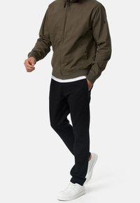 Veste bomber vert olive avec une fermeture éclair à l'avant, des poignets côtelés et deux poches latérales. Associée à un pantalon de jogging noir et des baskets blanches.