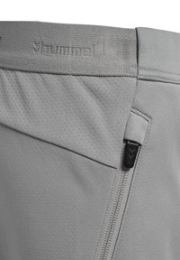 Close-up of gray athletic shorts waistband with textured fabric, embossed brand name "hummel," and vertical zipper pocket with black pull tab.