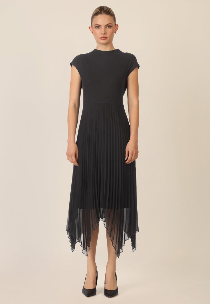 Vestido negro con un cuerpo ajustado y una falda plisada y fluida. Presenta un escote alto y un dobladillo asimétrico, hecho de una tela ligera.