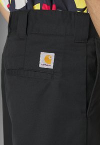 Sorte stofbukser med en glat tekstur, featuring en hvid lap med et gult logo og en detalje med en baglomme.