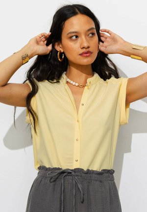 Femme aux longs cheveux noirs portant un chemisier jaune à manches courtes, un pantalon gris avec cordon de serrage, des boucles d’oreilles créoles dorées, un collier de perles et des bracelets dorés.