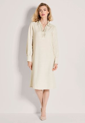 Vrouw draagt een beige midi-jurk met lange mouwen en een geborduurde kraag, gecombineerd met beige geweven pumps met puntige neus, staand tegen een effen achtergrond.