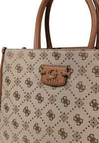 Beige tote bag with brown monogram pattern, leather handles, and metal logo accent. Features a structured design and patterned fabric.