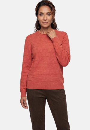 Woman with wavy hair wearing a textured long-sleeve coral sweater and dark brown pants, standing against a plain light background.