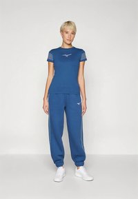 Γυναίκα με κοντά ξανθά μαλλιά, φορώντας μπλε αθλητικό μπλουζάκι και αντίστοιχες φόρμες, με λευκά αθλητικά παπούτσια, standing against a plain background.