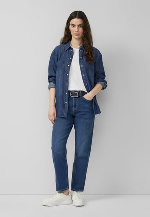 Femme debout portant une chemise en jean bleue sur un t-shirt blanc, un jean bleu, une ceinture noire et des baskets blanches devant un mur gris uni.
