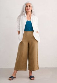 Cămașă albă din in, top teal fără mâneci și pantaloni largi din khaki. Sandale bleumarin cu detalii din curea țesută. Păr drept, gri.