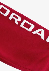 Tessuto rosso con una superficie liscia che presenta grandi lettere ricamate in bianco che scrivono "JORDAN". Il design è angolare e moderno.