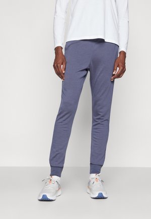 Mann trägt blaue Slim-Fit-Jogginghosen, weißes Langarmshirt und weiße Sportschuhe, steht vor einem schlichten Hintergrund.