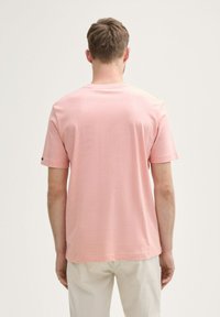 Mand, der vender væk, iført en lys pink kortærmet skjorte og lysebeige bukser foran en ensfarvet hvid baggrund.