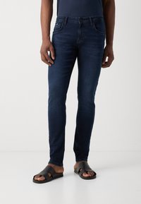 Tummansiniset slim fit -farkut, puuvillasekoitetta, etutaskut, hienovarainen haalistuminen, yhdistetty mustiin liukujalkineisiin, joissa on teksturoidut hihnat ja litteät pohjat.