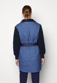 Blå quiltet lang trenchcoat med teksturerede marineblå ærmer, en blød krave og et stofbælte. Har et ensartet mønster og en afslappet pasform.