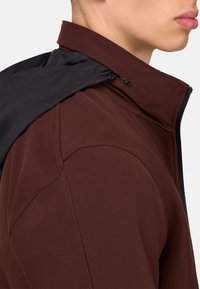 Hnědá zipová bunda s šedou kapucí. Má hladkou, strukturovanou tkaninu s jemnými švy a vysokým límcem. Navržena pro vysoký výkon.