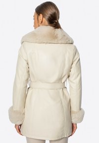 Kremowy płaszcz z syntetycznej skóry z szerokim futrzanym kołnierzem, dopasowanymi mankietami i paskiem do przewiązania. Posiada dopasowany krój oraz widoczne detale szwów.