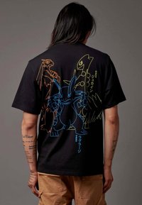 Zwarte T-shirt met kleurrijke cartoonontwerpen van Pokémon-personages op de achterkant, met blauwe, oranje en gele omtrek en tekst.