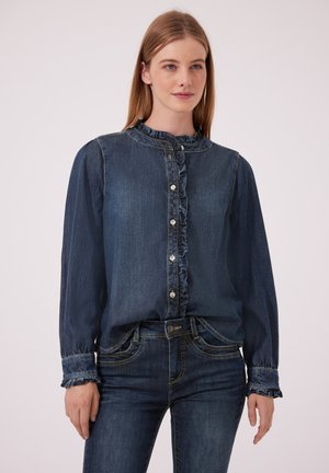 Vrouw die een donkerblauwe denim blouse met knopen en ruches draagt, samen met bijpassende donkerblauwe jeans, staand tegen een effen achtergrond.