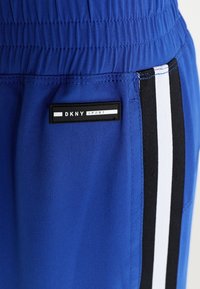 Blå sportshorts med svarta och vita sidostripes, elastisk midja och en svart etikett med texten "DKNY SPORT." Slät tygtextur.