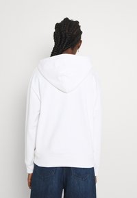 Persona con el cabello trenzado, vistiendo una sudadera blanca sencilla y jeans azules, de espaldas frente a una pared gris claro.