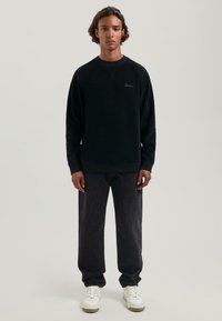 Joven de pie, recto, vestido con un sudadera negra, jeans negros holgados y zapatillas blancas, contra un fondo claro liso.