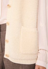 Cardigan crème, texturé avec une surface douce et duveteuse, doté d'une poche, d'un ourlet côtelé et de boutons. Porté sur un chemisier léger.