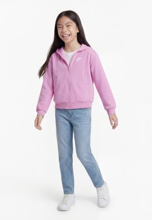 Nasmejana deklica z dolgimi lasmi, oblečena v roza Nike pulover s patentnim zobcem, svetlo modre kavbojke in bele superge, pozira na belem ozadju.