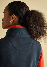 Tmavomodrý fleecový vest s oranžovým rebrovaným golierom, na chrbte s embosovaným nápisom "JOULES". Viditeľné kučeravé vlasy a strieborný krúžok v uchu.