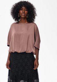 Vrouw met krullend haar, gekleed in gouden oorbellen, een losse mauve blouse en een zwarte kanten rok, staand tegen een effen witte achtergrond.