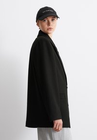 Černý oversized blazer s klopovým límcem a předními kapsami. Nositý přes šedý pruhovaný oblek. Doplněný černou kšiltovkou s bílým textem.