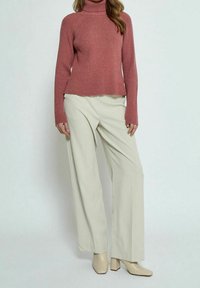 Pull à col montant côtelé rose clair associé à un pantalon large crème et des chaussures à talons carrés beiges, sur fond neutre.