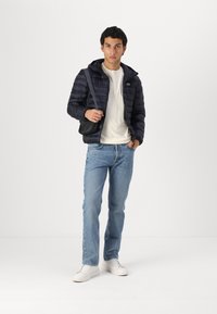 Marineblauwe pufferjack met capuchon, witte T-shirt, lichtblauwe denim jeans en witte sneakers, met een zwarte schoudertas in de hand.