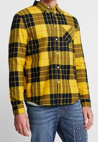 Żółto-czarna flanelowa koszula w kratę z przodem na guziki, pojedynczą kieszenią na piersi i długimi rękawami. Miękka w dotyku, z wzorzystym designem.