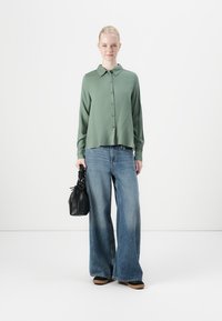 Chemise verte à manches longues avec col et boutons, associée à un jean en denim bleu à jambes larges. Le modèle tient un petit sac noir texturé.