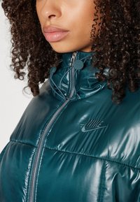 Tyrkysová nafukovacia bunda s lesklým povrchom, so s vysokým golierom, zipsom a vyšitým logom Nike na hrudi. Nastaviteľná šnúrka na golieri.