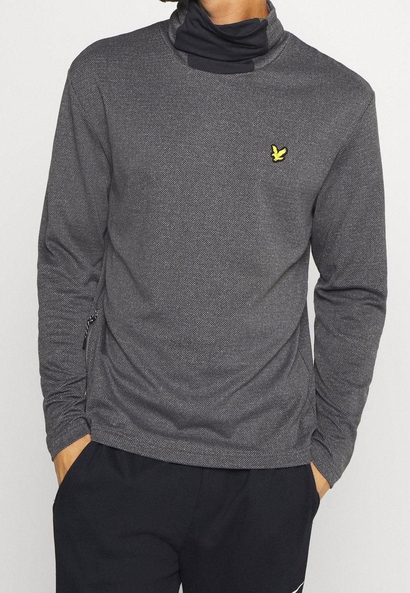 Haut gris à manches longues avec une finition texturée, doté d'un col montant et d'un petit logo jaune sur la poitrine. Comprend une poche zippée sur le côté.