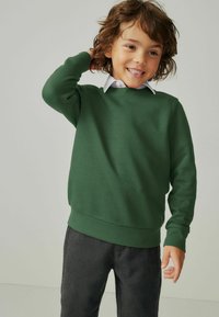Felpa verde in morbido tessuto con scollatura rotonda e polsini a coste, abbinata a una camicia bianca con colletto sotto e pantaloni grigi.
