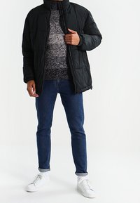 Schwarze Steppjacke mit hohem Kragen, kombiniert mit einem gemusterten Pullover, blauen Jeans und weißen Sneakers. Lässiges urbanes Outfit mit geschichteten Texturen.