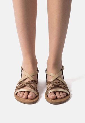 Füße in flachen Sandalen mit beige-braunen überkreuzten Riemen und Knöchelschnallen, stehend auf weißem Hintergrund.
