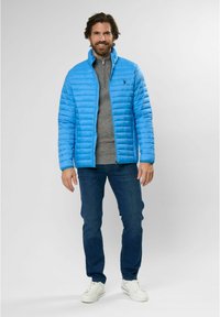 Svetlomodrá prešívaná bunda so zipsom, pod ňou šedý sveter a džínsové jeansy. Outfit doplňujú tenisky.