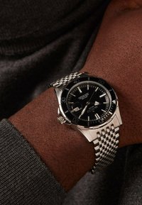 Silver metal wristwatch with a black dial, luminous hour markers, and a textured mesh bracelet. Features a date window and rotating bezel.