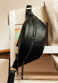 Borsa a tracolla in pelle nera dalla forma curva, con cerniere metalliche e tracolla testurizzata, esposta su una sedia rustica di legno.