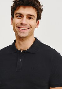 Czarny polo z teksturowanej tkaniny, z kołnierzykiem i zapięciem na dwa guziki, z wyszywanym logo na piersi.