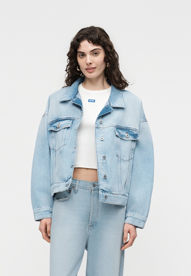 Femme aux cheveux bouclés et foncés portant une veste en denim bleu clair et un jean assorti par-dessus un top court blanc avec un petit logo bleu.