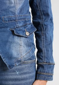 Jeansjacka i en urtvättad blå nyans med en texturerad yta, med en ficka med knäppning och kontrasterande sömmar.