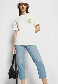 Camiseta branca de algodão com mangas curtas, apresentando um design gráfico colorido no lado esquerdo do peito. Combinada com jeans cropped de cor azul clara e uma mala preta.