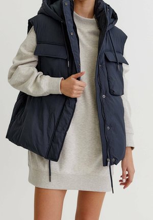 Frau trägt hellgraues Sweatshirtkleid mit langen Ärmeln und eine offene marineblaue gesteppte Weste mit Klappentaschen und Kordeln.