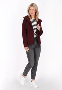 Burgundska jakna z zadrgo z visokim ovratnikom, nošena preko črno-belega črtastega srajca, kombinirana z sivimi jeans hlačami in belimi športnimi copati.