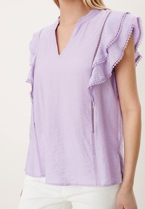 Kobieta ubrana w lawendową bluzkę bez rękawów z marszczonymi rękawami wykończonymi koronką i dekoltem w serek, zestawioną z białymi spodniami.