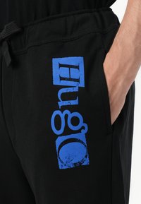 Fekete jogger nadrág, amelyen egy nagy kék "Hugo" logó található, strukturált felülettel a bal lábon. Rugalmas derékpánt, húzózsinórral.